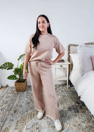 Woman in a beige outfit standing in a bedroom with a plant and bed in the background