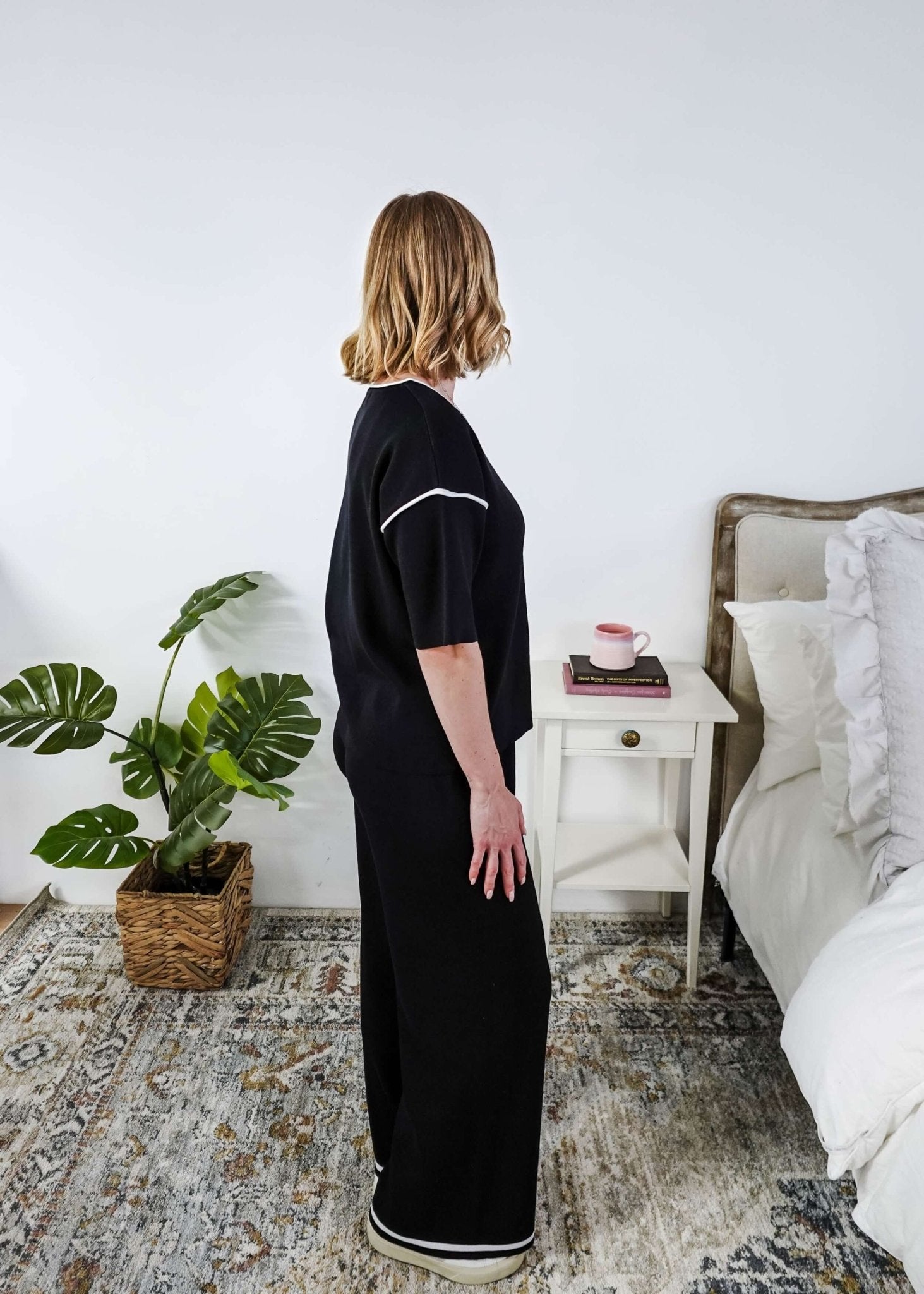 Woman in a black lounge outfit standing in a bedroom with a plant and bed in the background