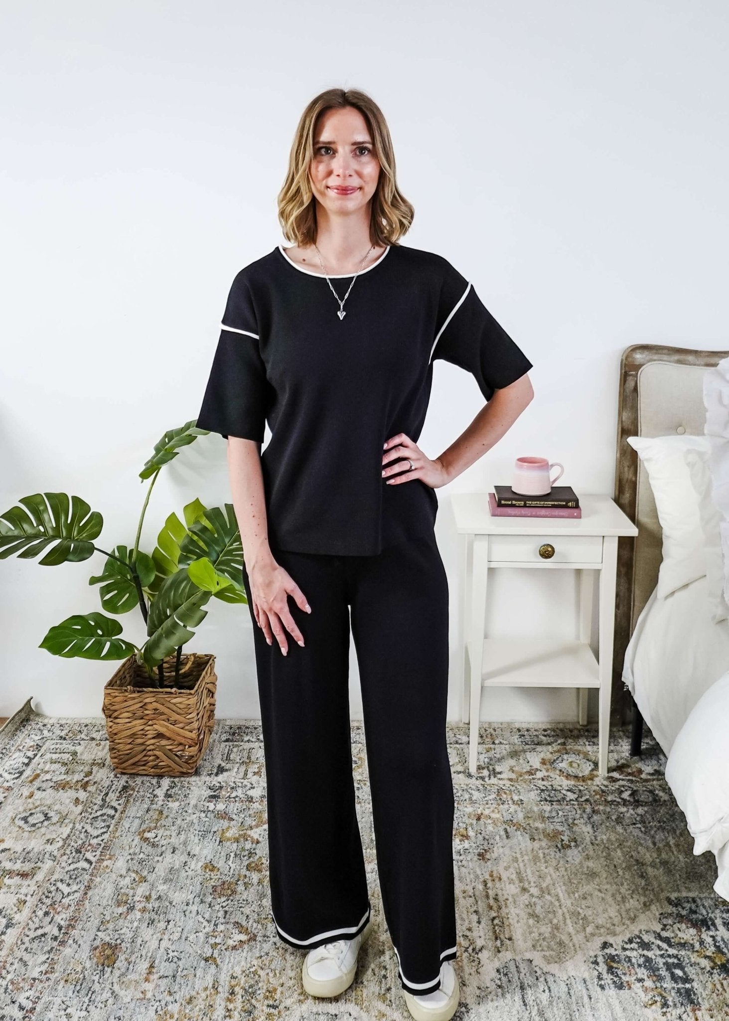 Woman wearing a black outfit in a bedroom setting with a plant and bed in the background.