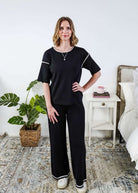 Woman wearing a black outfit in a bedroom setting with a plant and bed in the background.