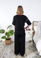 Person wearing a black lounge outfit standing in a bedroom with a plant and nightstand.