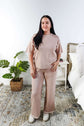 Woman wearing a beige outfit standing in a bedroom with a plant and bed in the background.