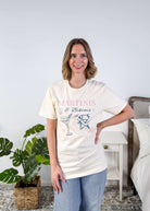 Martinis & Bikinis Relaxed Fit Graphic Tee with martini and bikini print in pink and blue and some seashells, worn on model with jeans in a bedroom setting