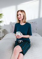 Midnight Plaid Unisex Bamboo Sleep Shorts worn by a woman sitting on a bed, holding a cup in a bright bedroom.
