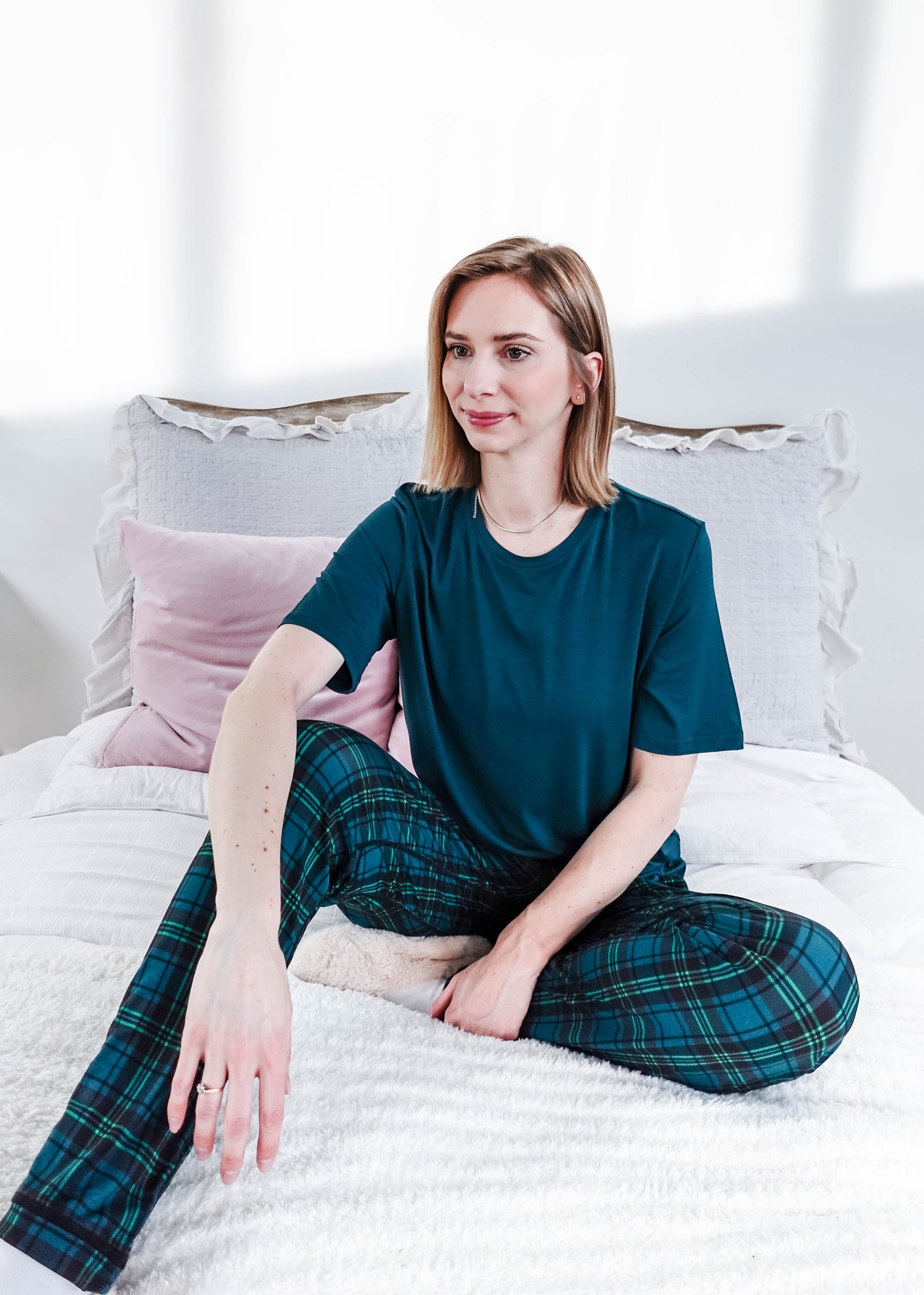 Model wearing Midnight Plaid Unisex Bamboo Joggers and a teal shirt, sitting on a bed with soft pillows.