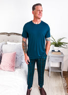 Model wearing Midnight Plaid Unisex Bamboo Joggers paired with a blue shirt in a cozy bedroom setting.
