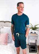 Man wearing Midnight Plaid Unisex Bamboo Sleep Shorts and a blue T-shirt in a cozy bedroom setting.