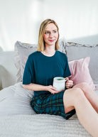 A woman sitting on a bed in Midnight Plaid Unisex Bamboo Sleep Shorts, holding a mug, enjoying a cozy atmosphere.