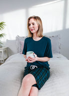 Midnight Plaid Unisex Bamboo Sleep Shorts worn by a woman sitting on a bed, holding a cup in a bright bedroom.