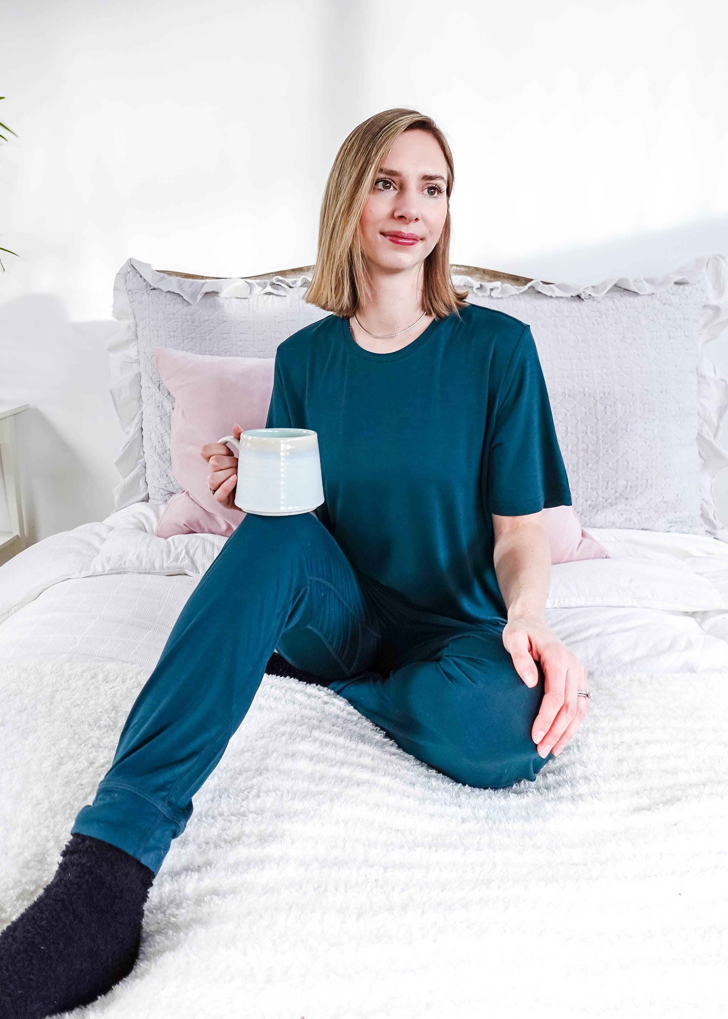 Model wearing Midnight Teal Unisex Bamboo Joggers while holding a cup and sitting on a bed.