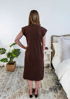 The back of a woman in a brown midi dress holding a beige handbag in a bedroom setting.
