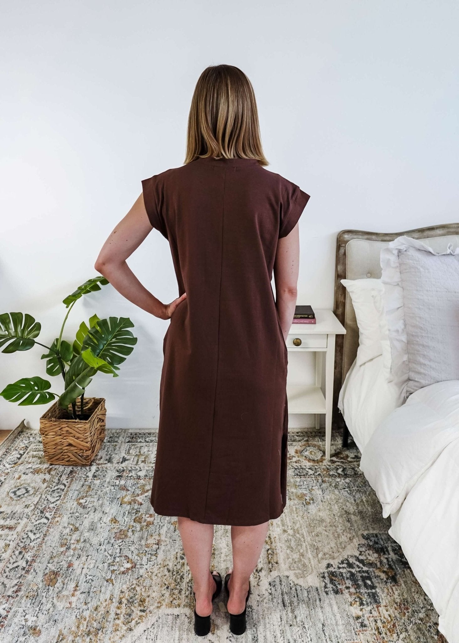 The back of a woman in a brown midi dress holding a beige handbag in a bedroom setting.