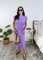 Woman in a purple  midi dress holding a beige handbag in a bedroom setting.