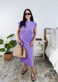 Woman in a purple  midi dress holding a beige handbag in a bedroom setting.