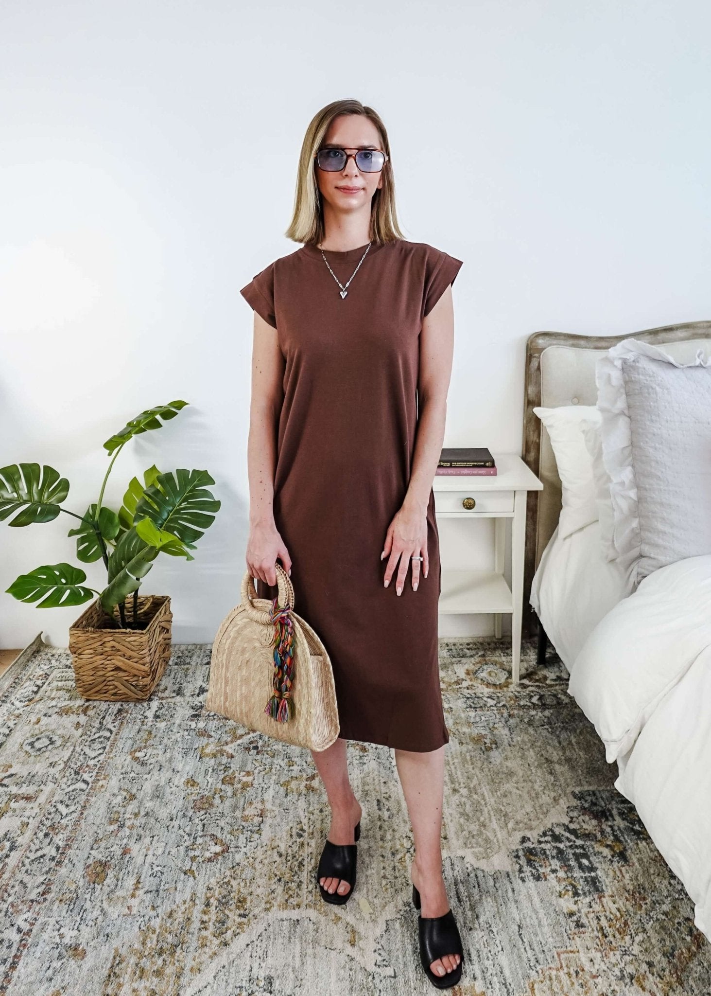 Woman in a brown midi dress holding a beige handbag in a bedroom setting.