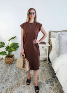 Woman in a brown midi dress holding a beige handbag in a bedroom setting.