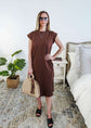 Woman in a brown midi dress holding a beige handbag in a bedroom setting.
