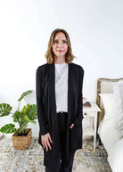 Woman in a black cardigan standing in a room with a plant and a bed in the background Scarlett Ellie Summer Open Lightweight Cardigan | Cloud Nine Pajamas