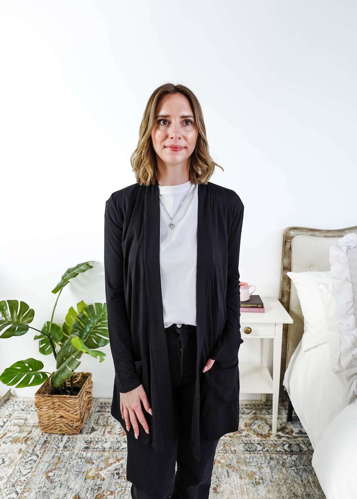 Woman in a black cardigan standing in a room with a plant and a bed in the background Scarlett Ellie Summer Open Lightweight Cardigan | Cloud Nine Pajamas