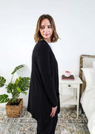 Woman in a black cardigan standing in a room with a plant and a bed in the background Scarlett Ellie Summer Open Lightweight Cardigan | Cloud Nine Pajamas