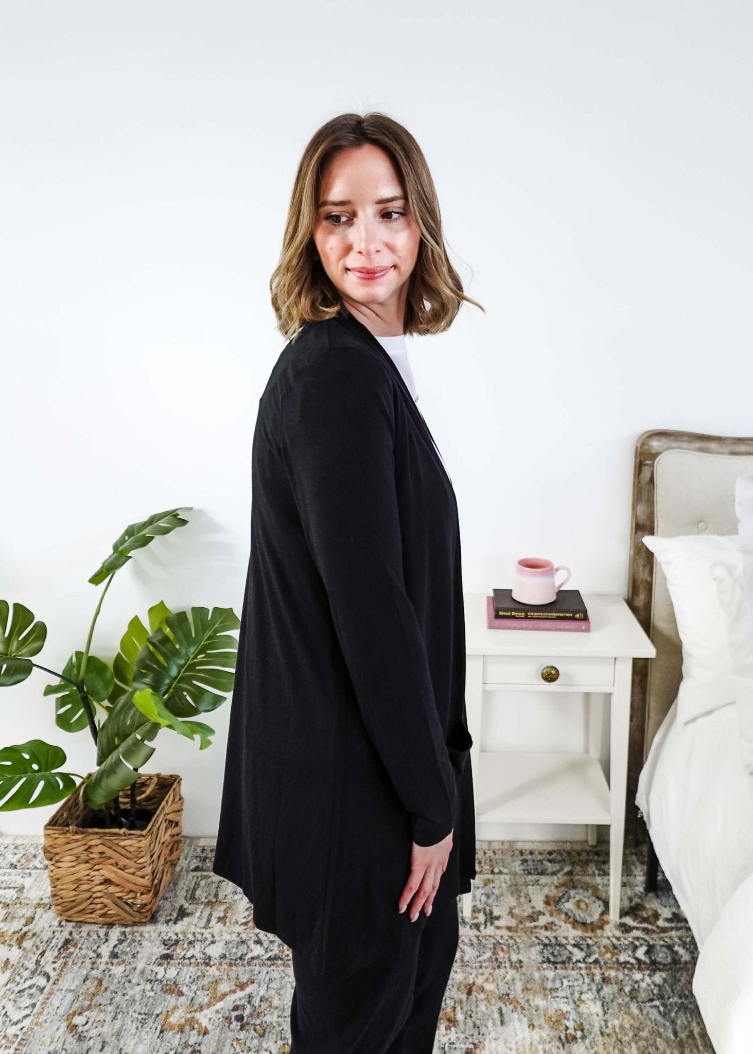 Woman in a black cardigan standing in a room with a plant and a bed in the background Scarlett Ellie Summer Open Lightweight Cardigan | Cloud Nine Pajamas