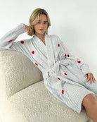 Model wearing the Pomona Fuzzy Yarn Robe Splendid in gray with red hearts, sitting on a plush chair.