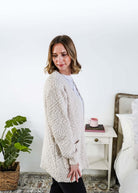Woman wearing a beige cardigan with texture  in a bedroom setting - Popcorn Texture Open Summer Cardigan showing side profile