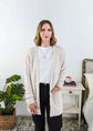 Popcorn Texture Open Summer Cardigan in sand beige, worn by Woman wearing a beige cardigan in a bedroom setting with a white tee and black jeans