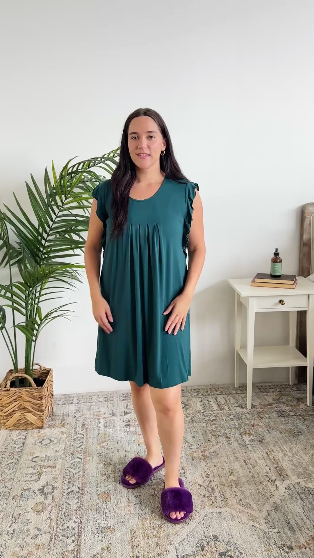 Women standing in bedroom showing her nightgown off. The night dress is dark teal with a flowy fit and pleating on the front. 