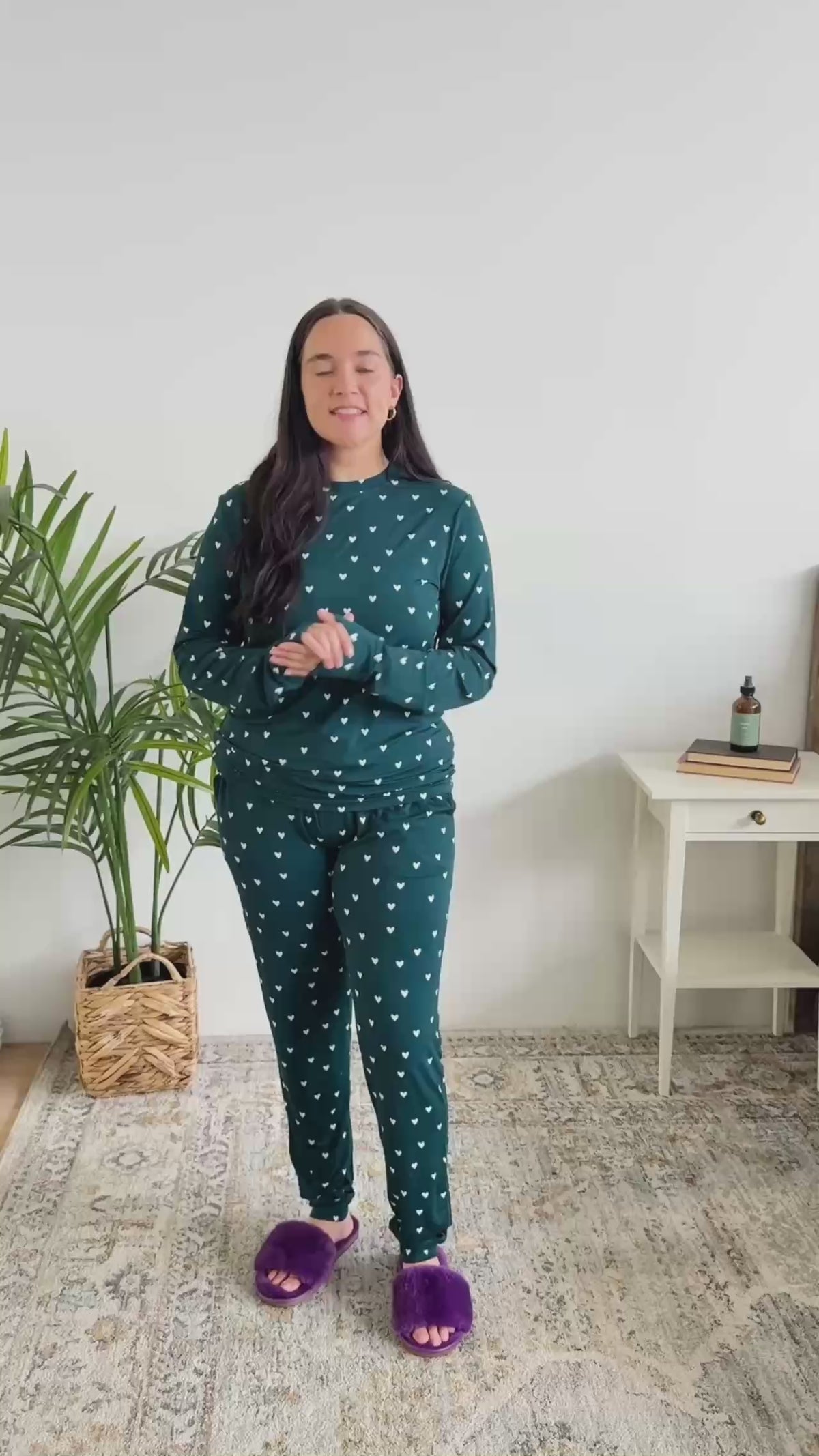 Women standing in bedroom showing her pajamas. The pajamas are a long sleeve top with long pants. The colour is dark teal with cute little white hearts. 