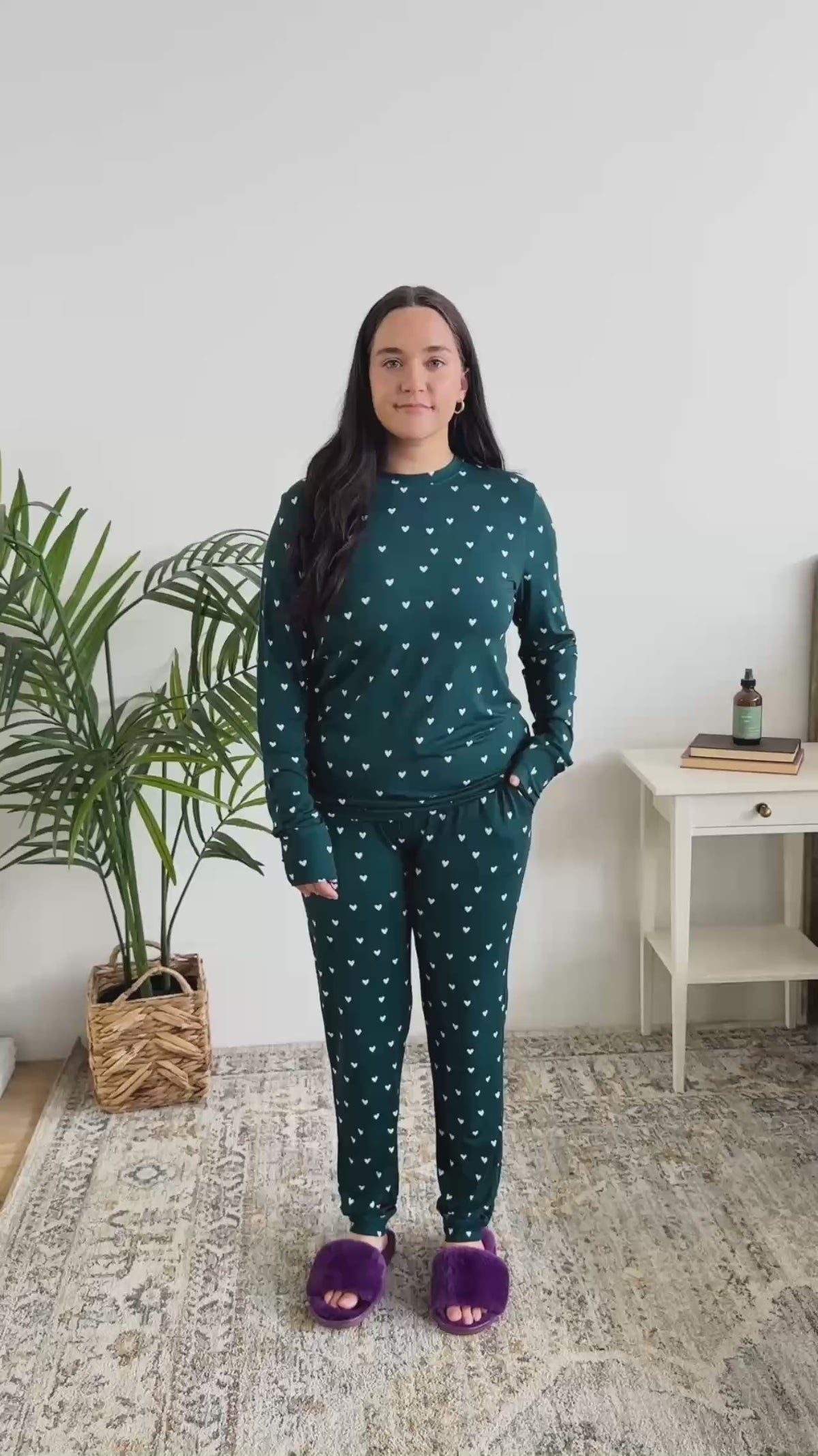 Women standing in bedroom showing her pajamas. The pajamas are a long sleeve top with long pants. The colour is dark teal with cute little white hearts. 