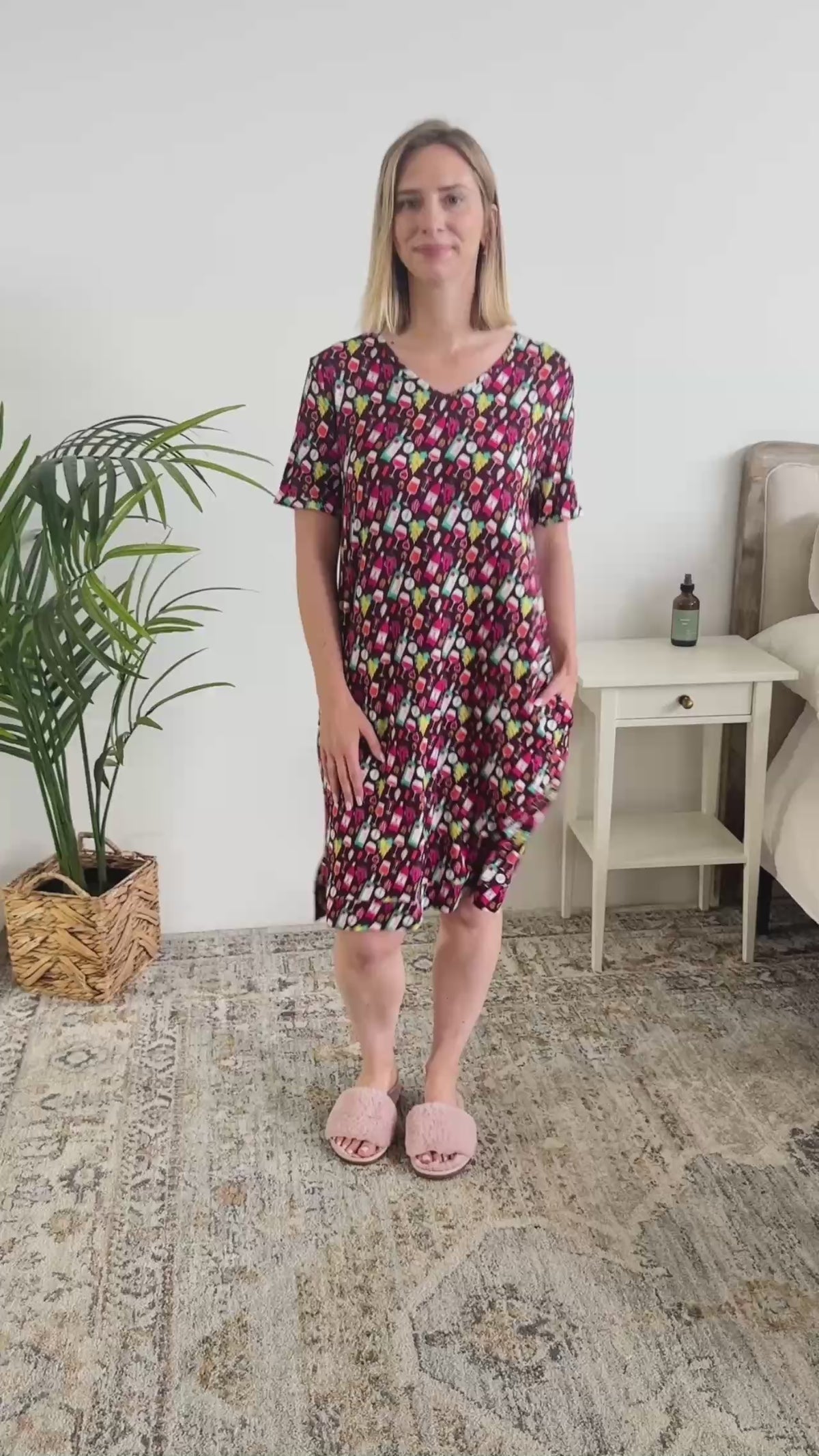 Women in a bedroom showing her nightgown. The night dress is knee length v-neck with a fun wine print. 