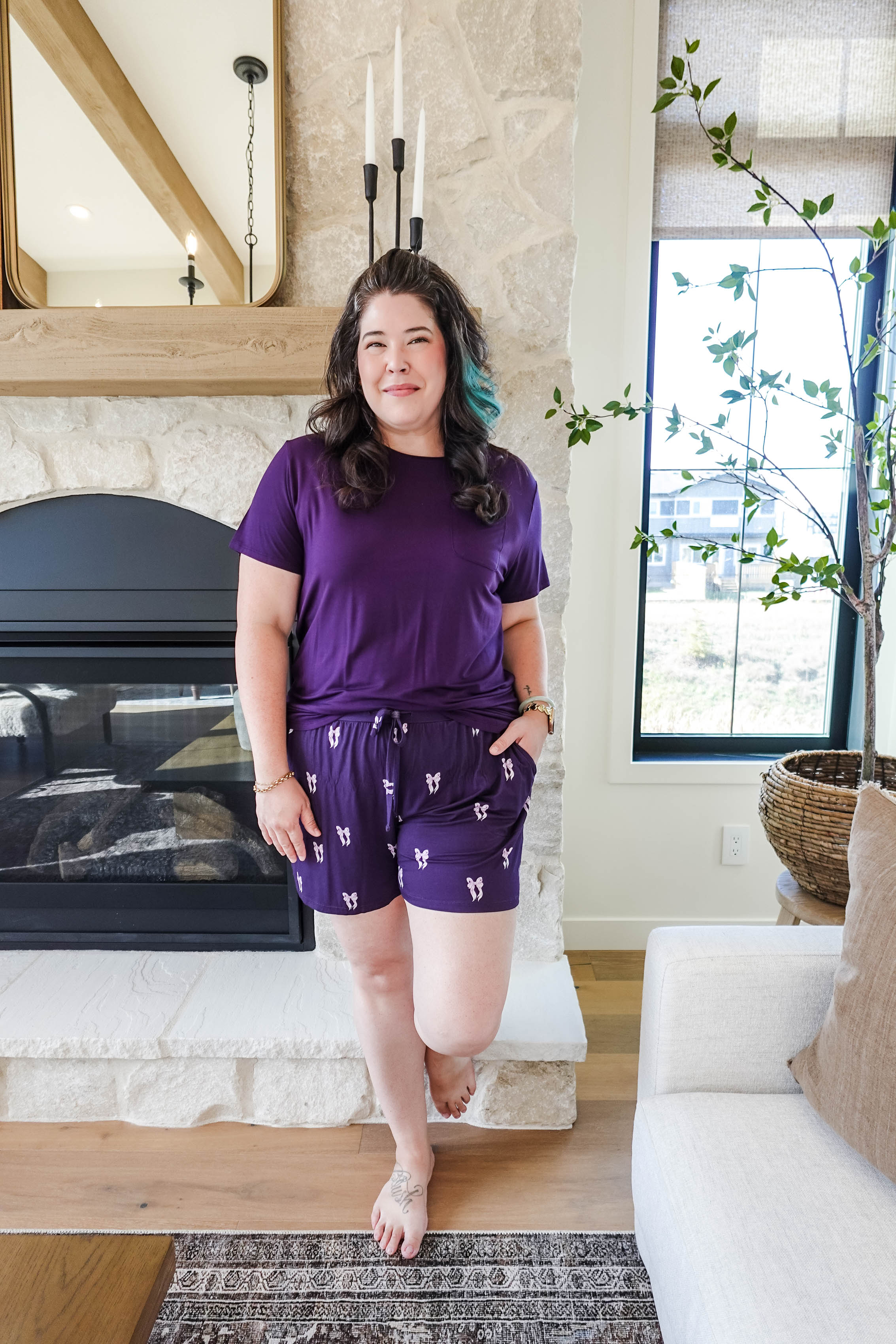 Woman wearing a Purple Bows Print Tee & Short Bamboo PJ Set by GEORGIA RYLAN, standing by a cozy fireplace.