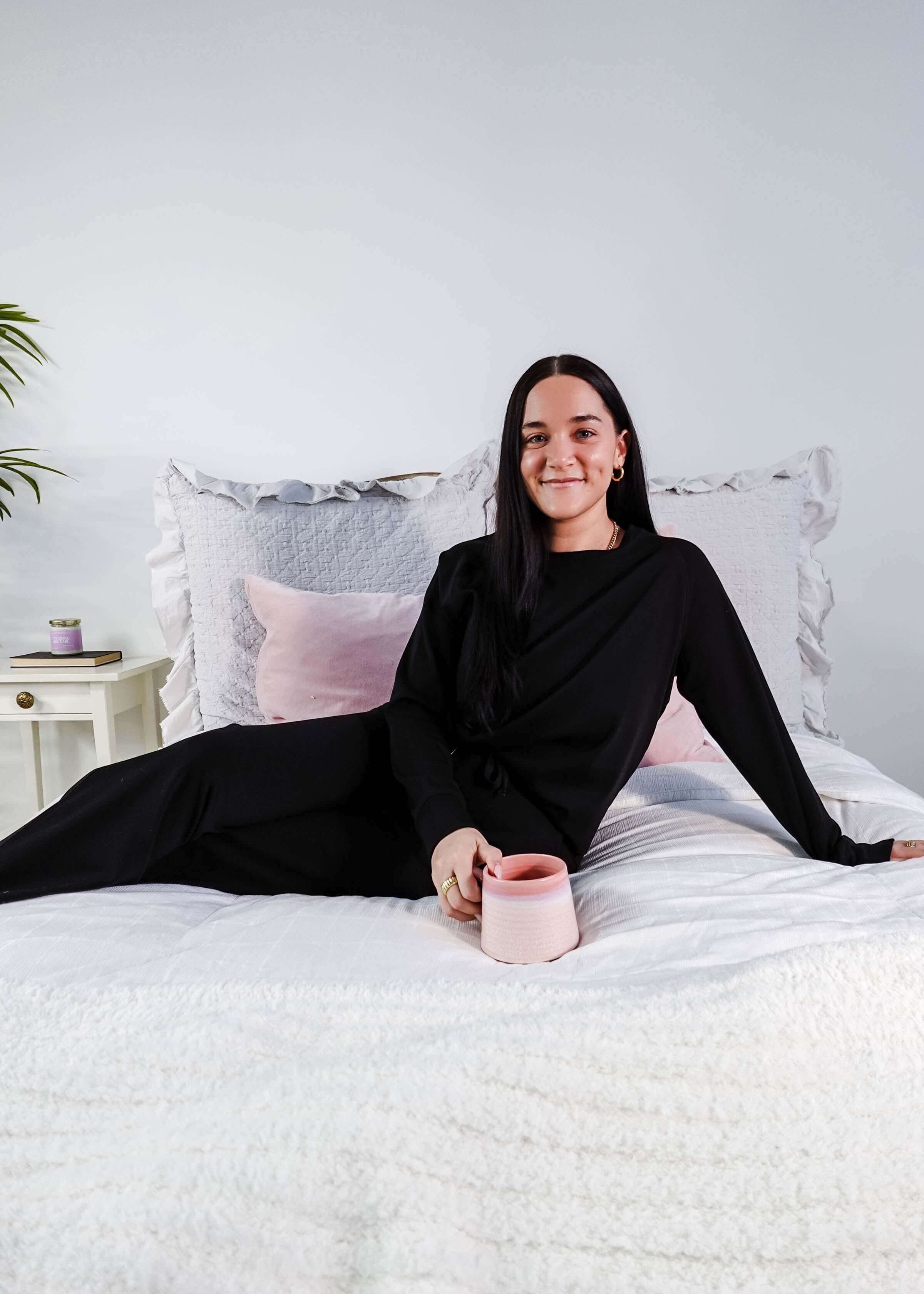 Model wearing Black Long Sleeve Raw Edge Straight Pant Cloud Lounge Set on a bed with a pink mug.