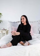 Woman wearing Black Long Sleeve Raw Edge Straight Pant Cloud Lounge Set seated on a bed holding a drink.