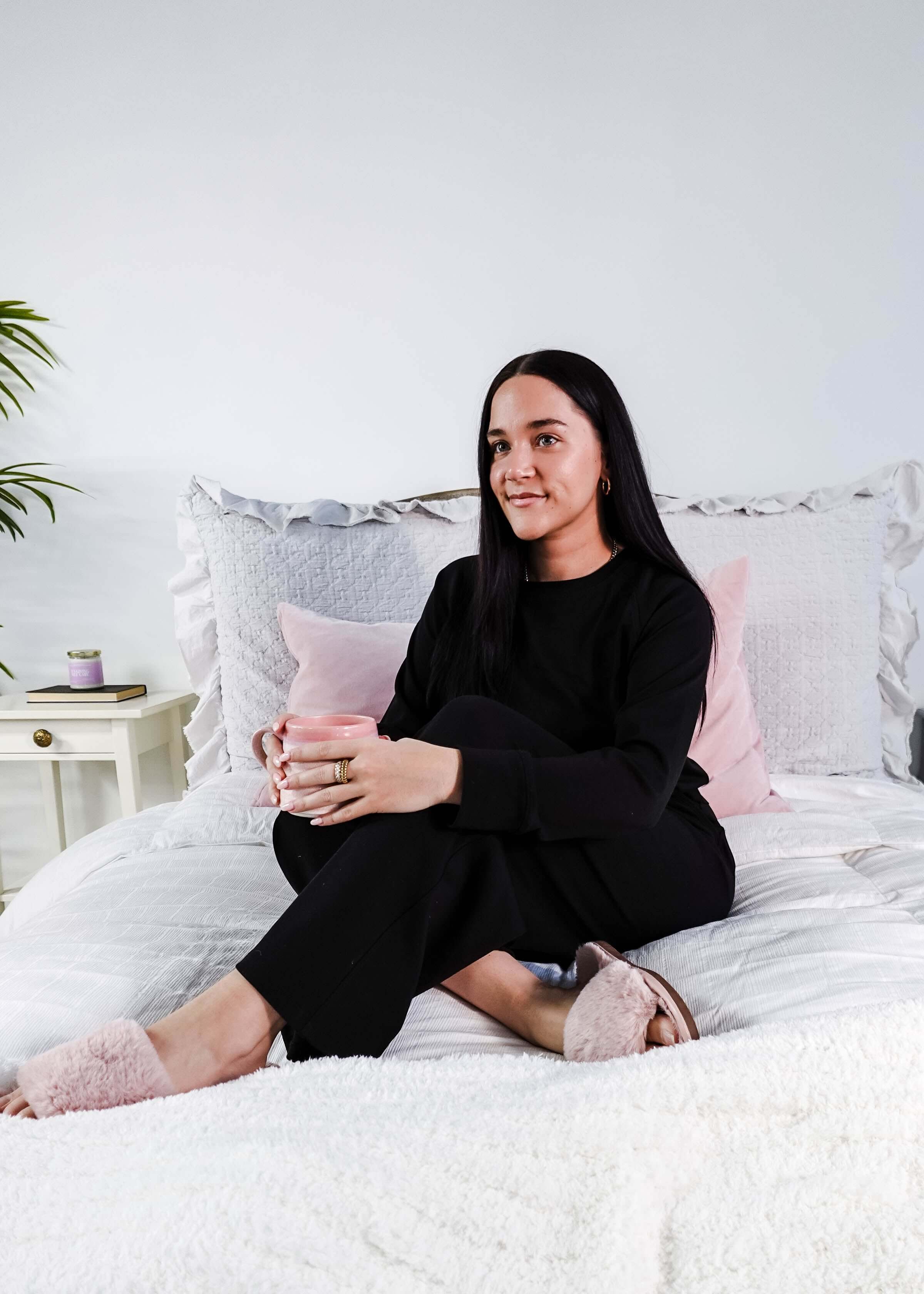 Woman wearing Black Long Sleeve Raw Edge Straight Pant Cloud Lounge Set seated on a bed holding a drink.