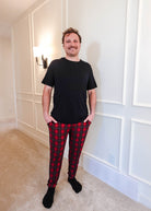 A man wearing a black shirt and Red Plaid Unisex Bamboo Jogger from Bob's Sleepwear Co. standing in a well-lit room.