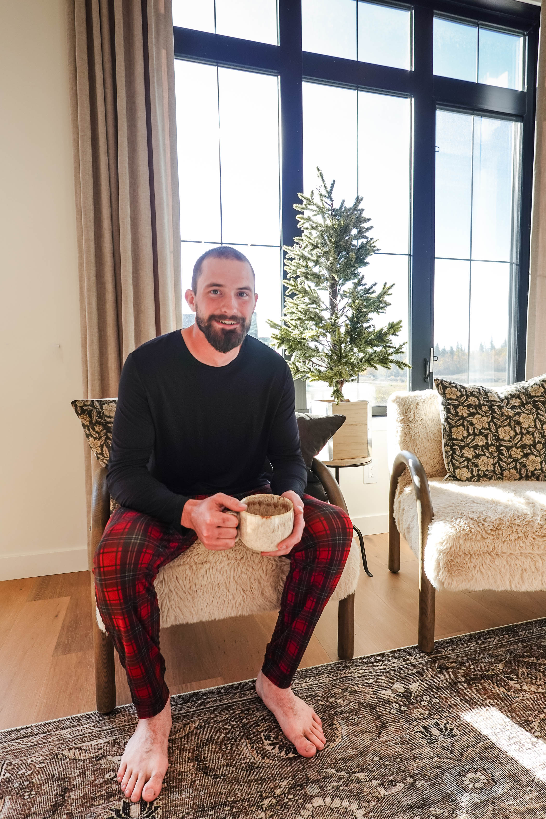 A man wearing Red Plaid Unisex Bamboo Jogger from Bob's Sleepwear Co. sitting with a mug in cozy interior.