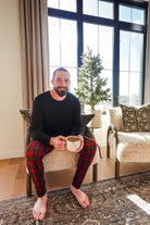 A man wearing Red Plaid Unisex Bamboo Jogger from Bob's Sleepwear Co. sitting with a mug in cozy interior.