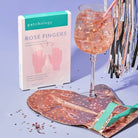 Patchology Rosé Fingers product with a glass of rosé and a patterned bag on a blue background, Patchology Rosé Fingers renewing hand mask rose gold gloves shea butter