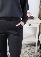 Person wearing black pants with a drawstring waistband, blurred background of a white shelf and books.