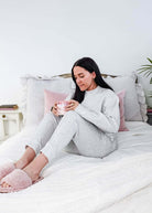 Woman in gray pajamas sitting on a bed holding a pink cup, surrounded by pillows and a plant.