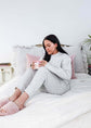 Woman in gray pajamas sitting on a bed holding a pink cup, surrounded by pillows and a plant.