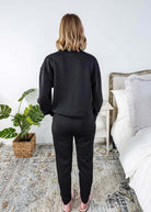 Person wearing a black outfit standing in a bedroom with a plant and bed in the background.