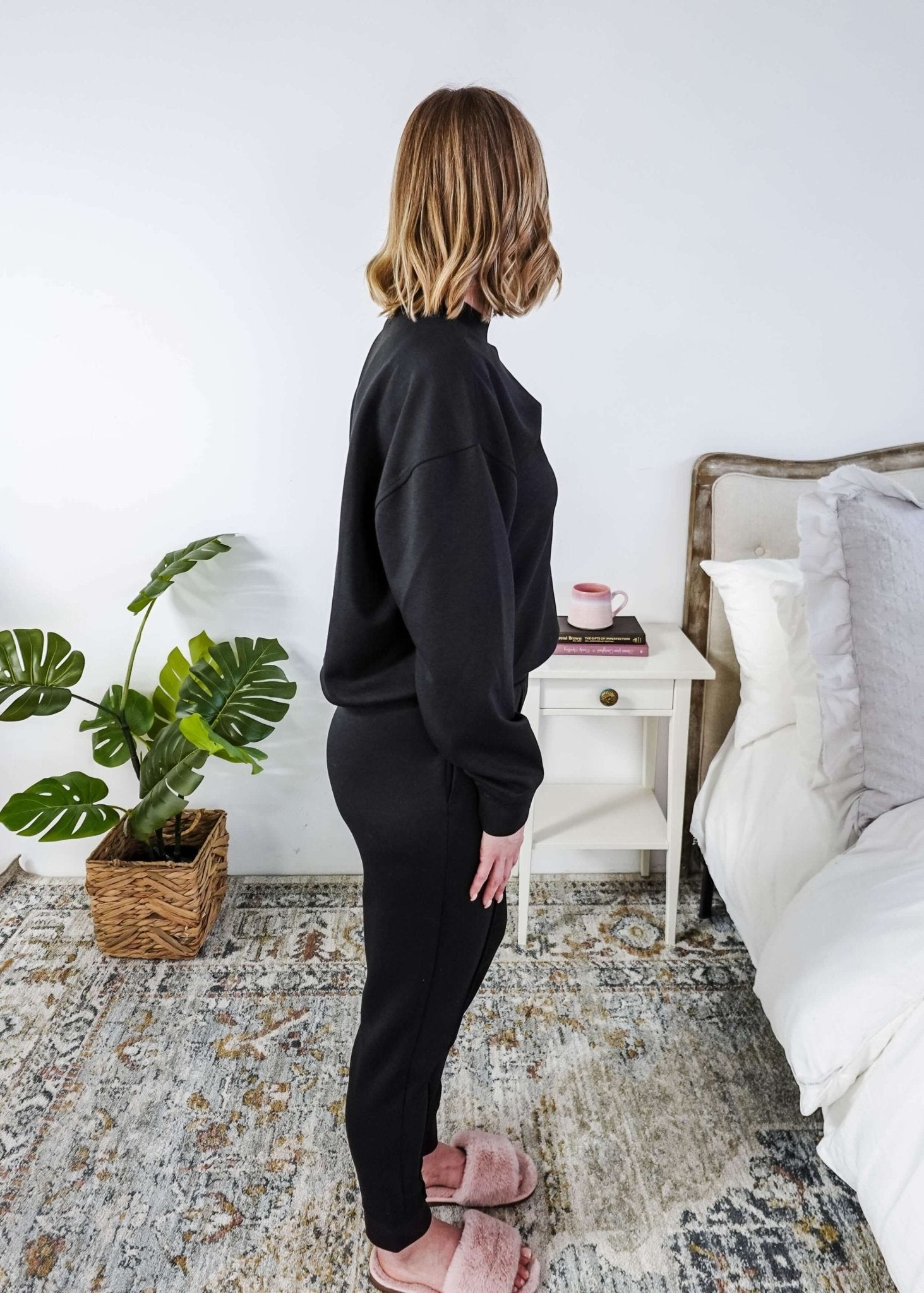 Woman in black tracksuit standing in a bedroom with a plant and bed in the background