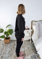 Woman in black tracksuit standing in a bedroom with a plant and bed in the background