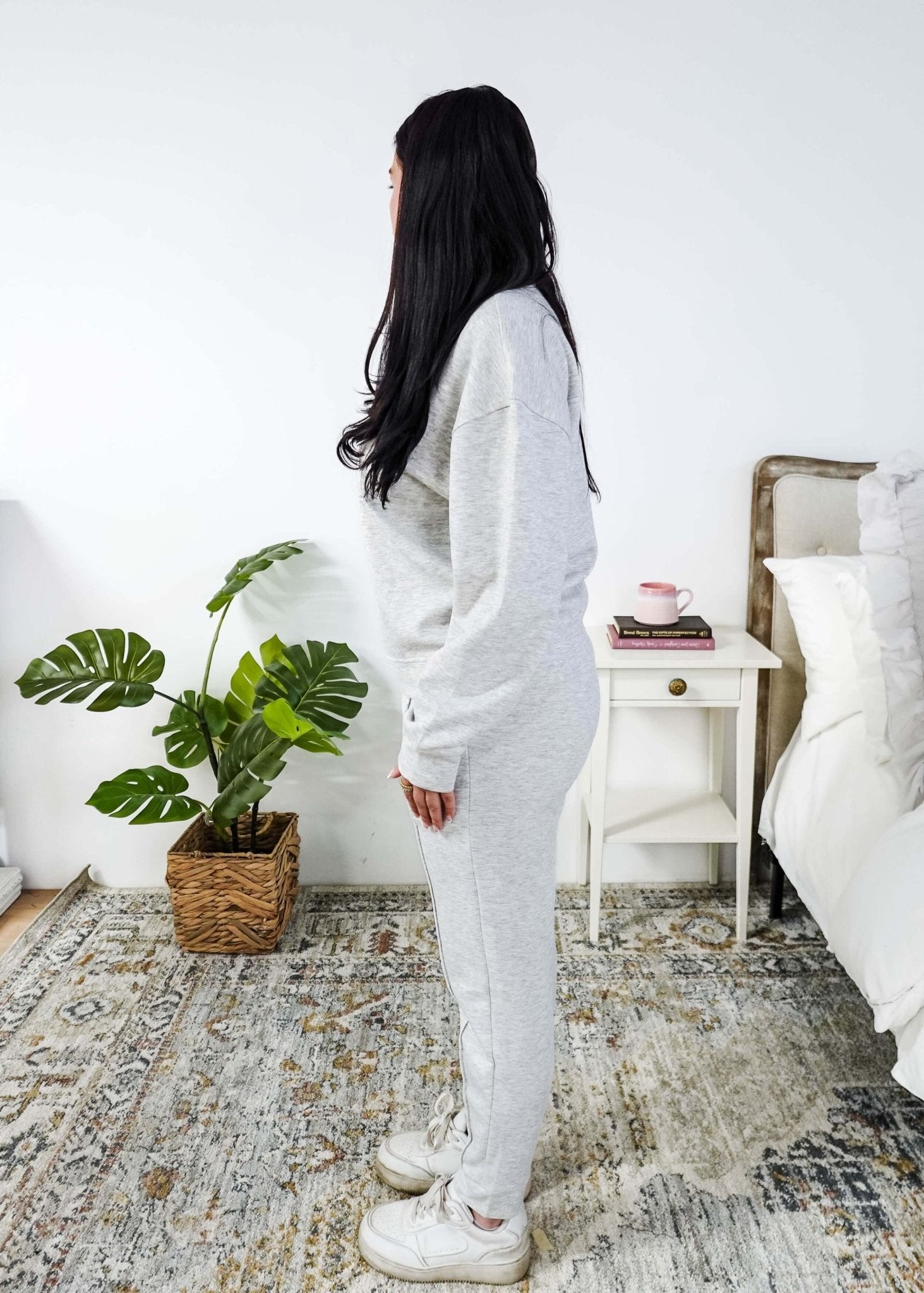 Woman in a cozy room with a plant and a nightstand wearing a grey lounge set. 