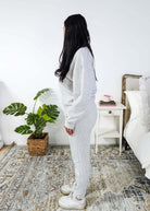 Woman in a cozy room with a plant and a nightstand wearing a grey lounge set. 