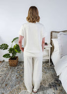 The back of a woman in a cream lounge outfit standing in a bedroom with a plant and bed in the background