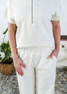 Woman in a cream lounge outfit standing in a bedroom with a plant and bed in the background. Pants are an elastic pnt. 